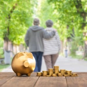 Cofrinho dourado e pilhas de moedas em primeiro plano, com um casal de idosos caminhando ao fundo, simbolizando planejamento financeiro para a aposentadoria.