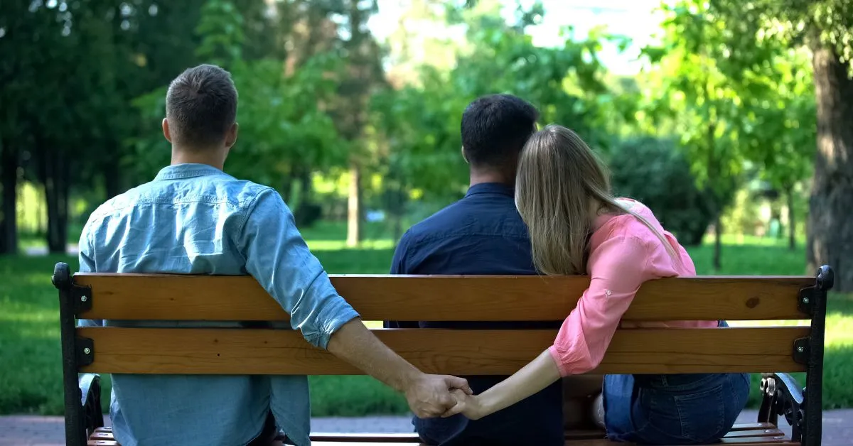 Homem e mulher sentados com outro homem em um banco de parque, enquanto se dão as mãos escondido pelas costas dele