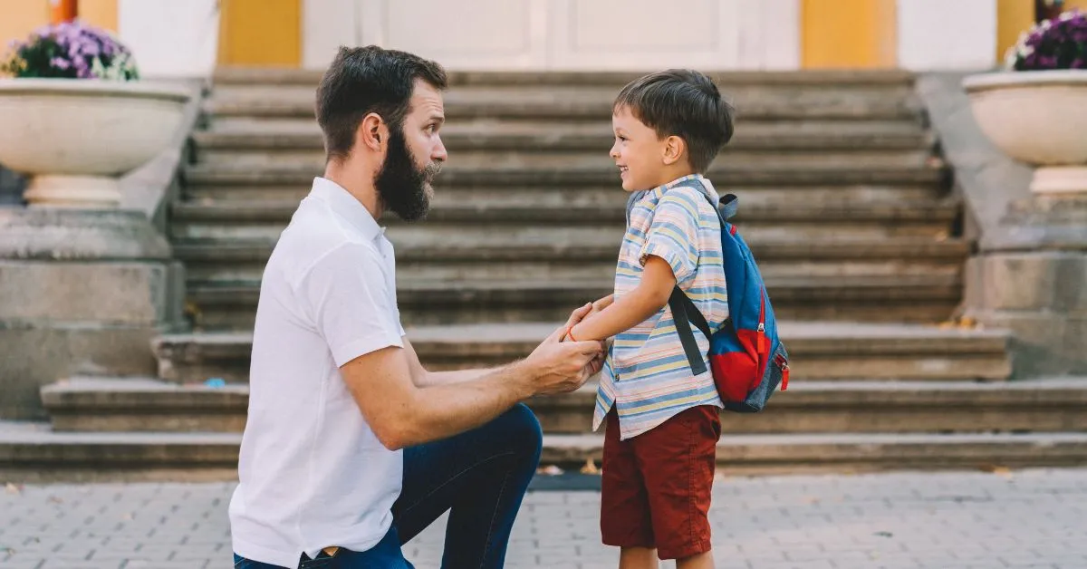 Alienação Parental Invertida: pai ajoelhado segura as mãos do filho com mochila em frente à escola