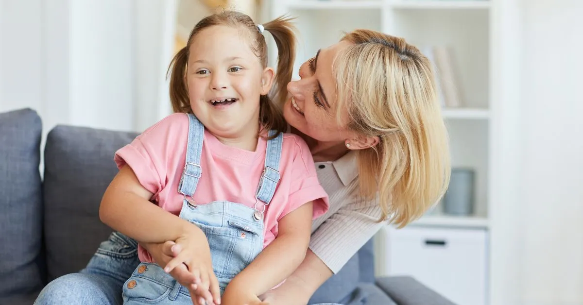 Adoção de criança com deficiência: mãe abraça filha sorridente no sofá, representando acolhimento, afeto e guarda responsável.